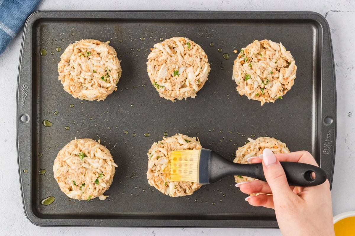 A hand brushing the tops of unbaked crab cakes with melted butter on a baking sheet.
