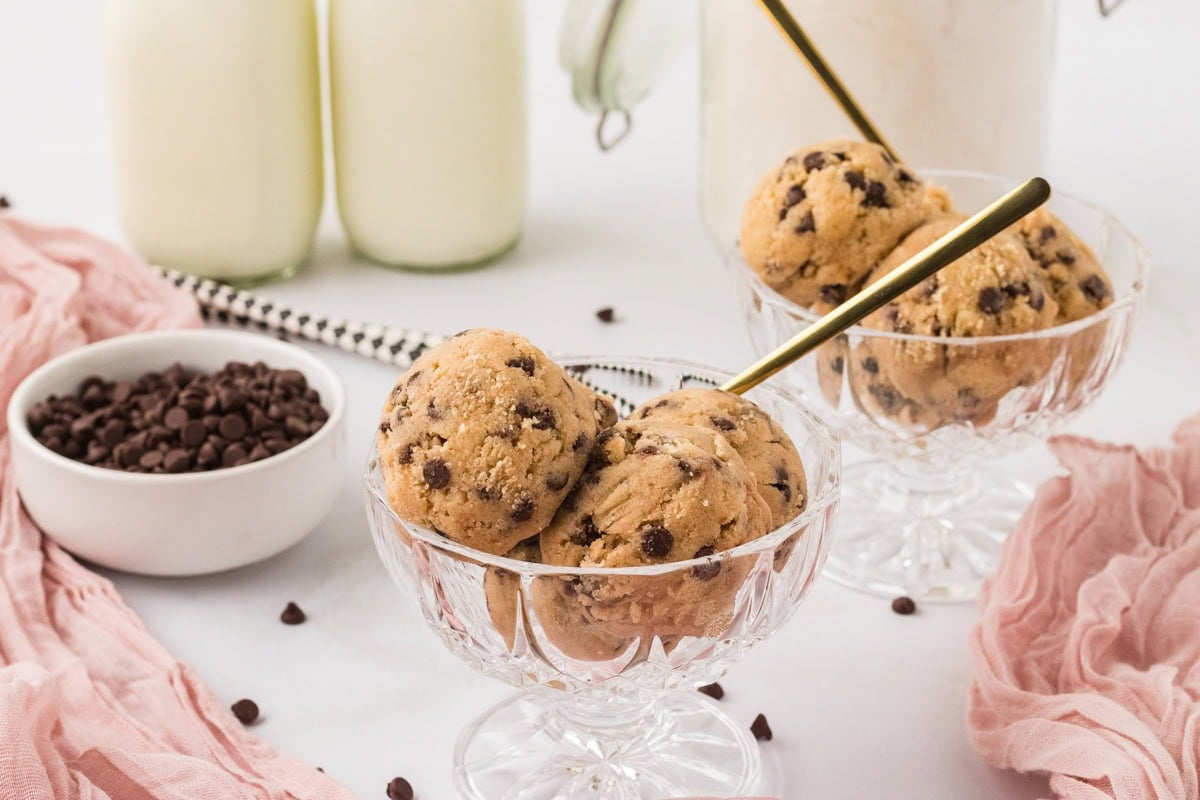 Glass dessert cups filled with scoops of gluten-free edible cookie dough, with mini chocolate chips and glasses of milk in the background.