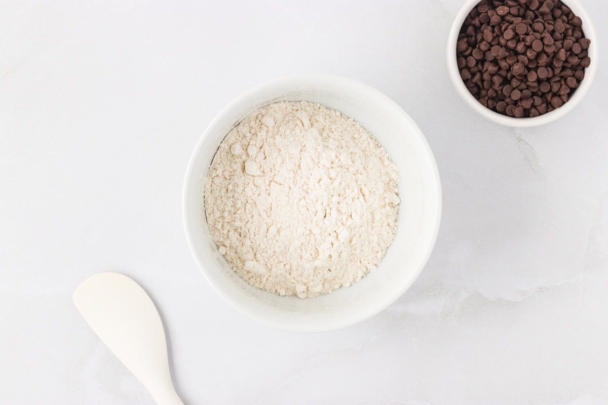 White bowl of gluten-free flour next to a small bowl of mini chocolate chips on a white surface with a spatula.
