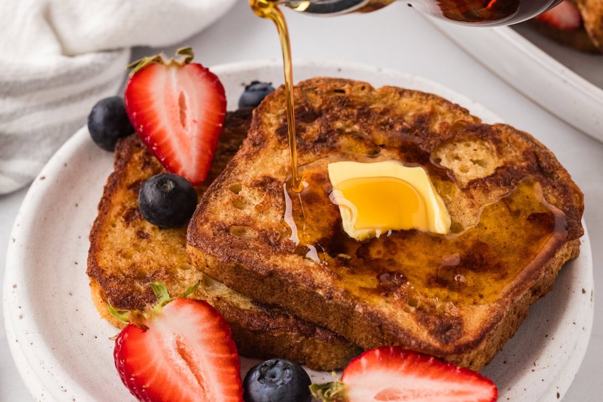 Close-up of French toast on a plate with a pat of melting butter and syrup being poured over the top, served with fresh berries.