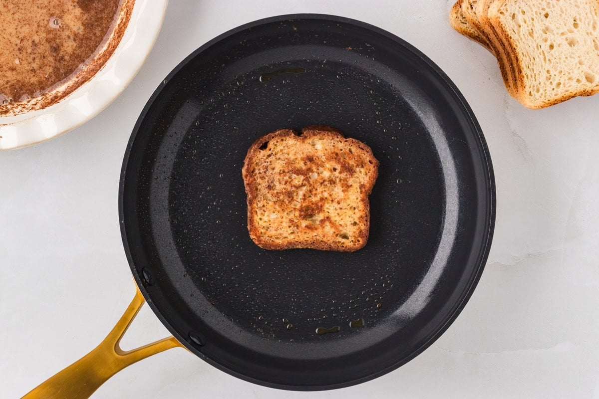 A single slice of gluten-free French toast cooking in a nonstick skillet, starting to brown on one side.