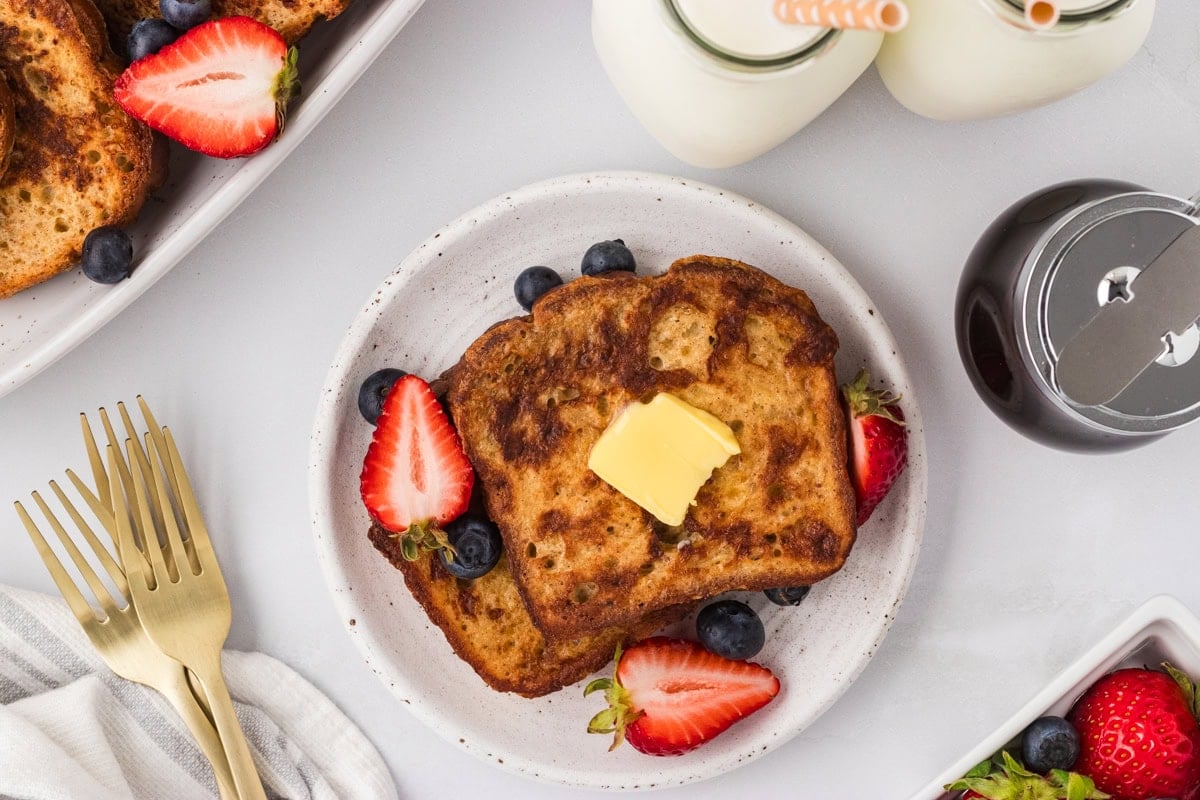 Plate of cooked gluten-free French toast topped with butter and surrounded by fresh strawberries and blueberries, with milk and syrup in the background.