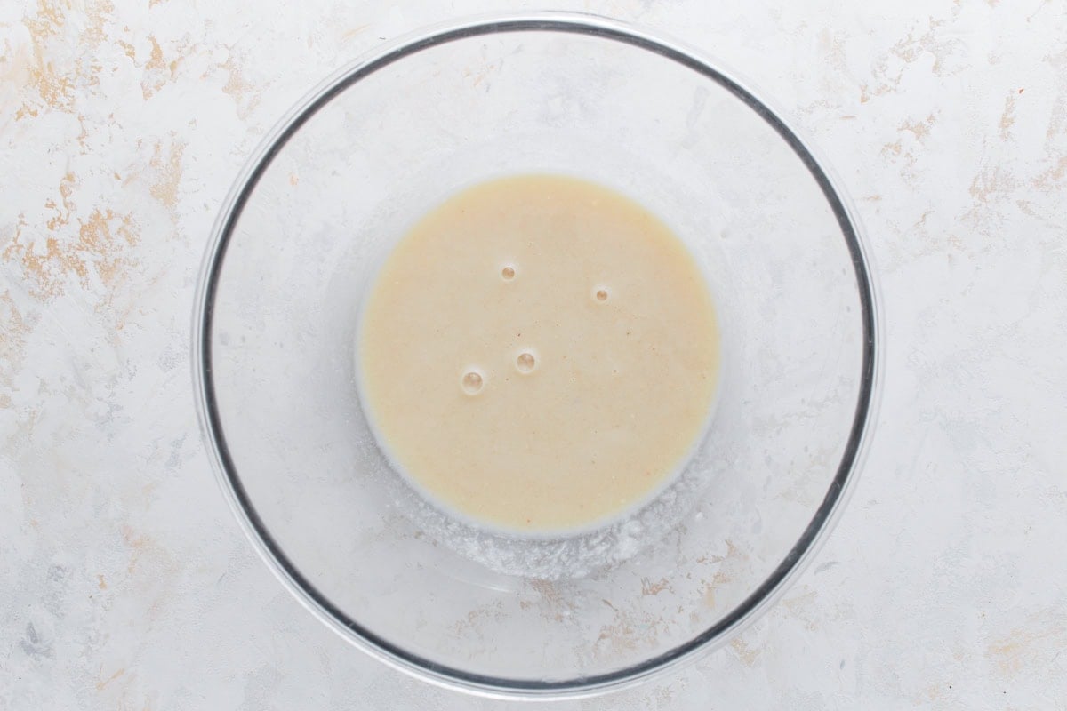 Clear mixing bowl with a smooth, beige liquid mixture of wet ingredients for oatmeal bars on a light textured background.