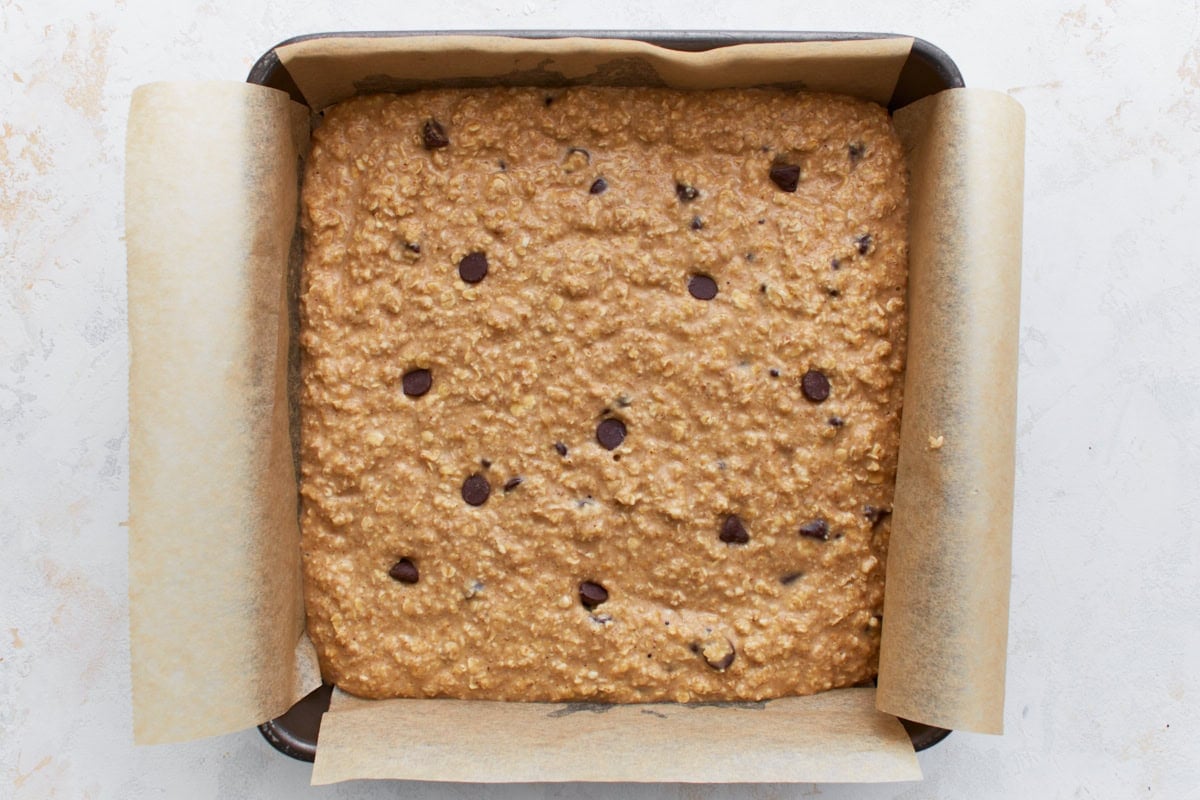Chocolate chip oatmeal batter spread evenly in a parchment-lined square baking pan before baking.