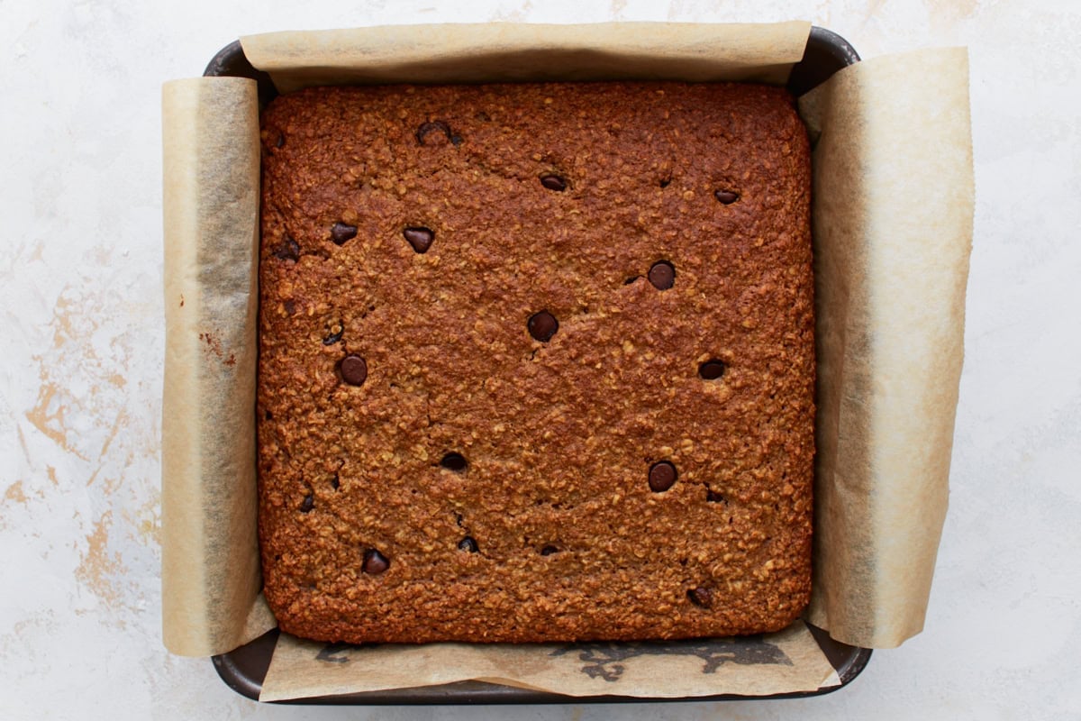 Fully baked oatmeal bar slab, golden brown and studded with melted chocolate chips, still in the parchment-lined pan.
