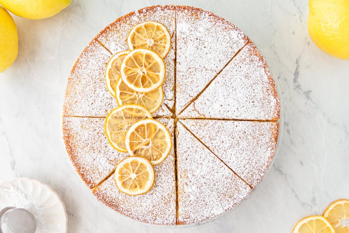Overhead view of a sliced gluten-free olive oil cake topped with powdered sugar and candied lemon slices, surrounded by fresh lemons.