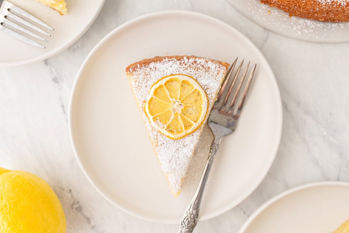 Single slice of gluten-free olive oil cake on a white plate, topped with powdered sugar and a lemon slice, with a fork beside it.