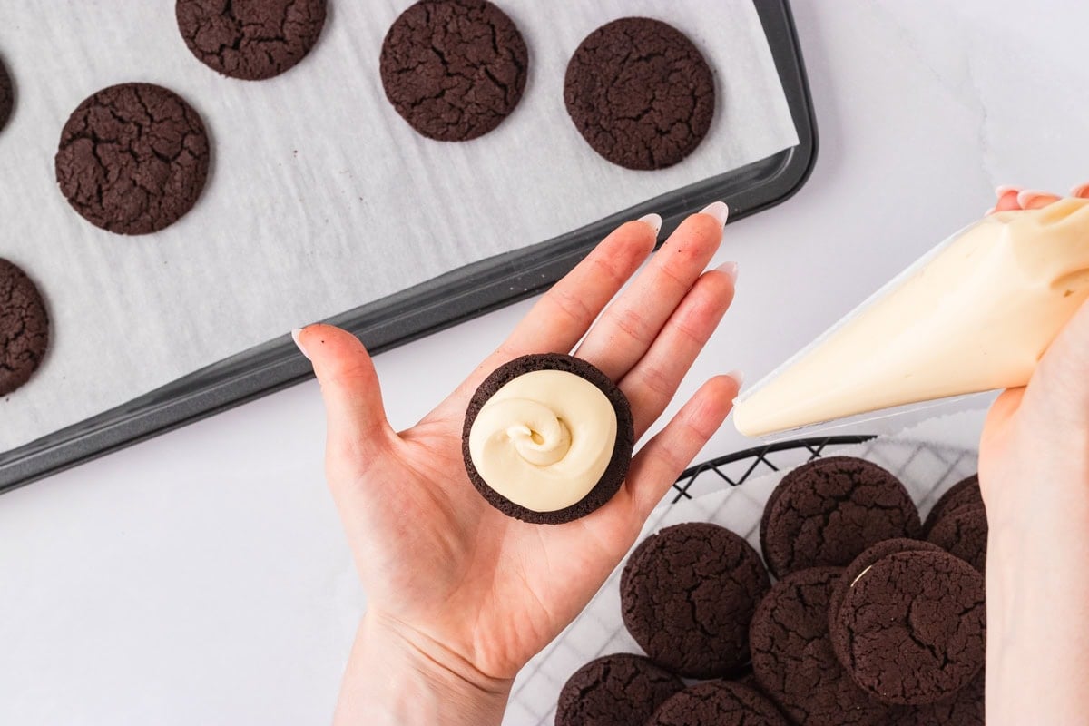 A hand holding a chocolate cookie with piped vanilla filling, using a piping bag to prepare the sandwich cookies on a cooling rack.