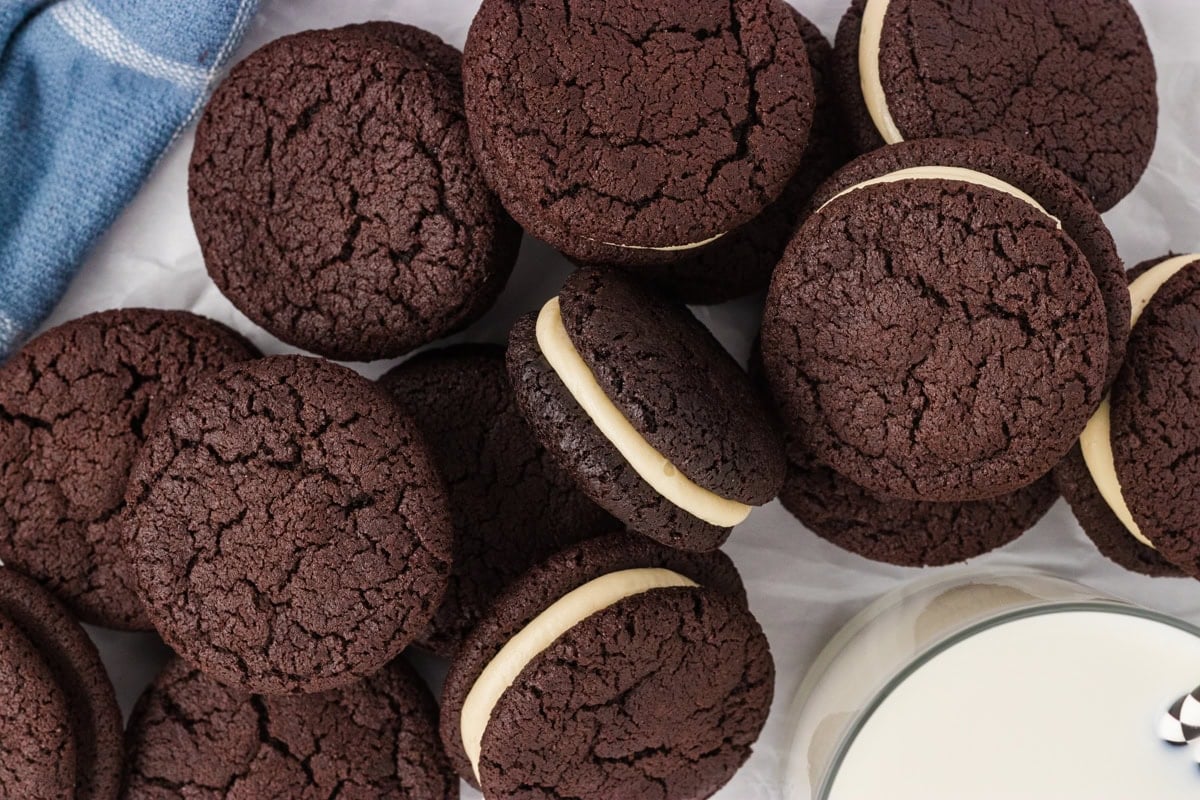 A close-up, top-down view of several gluten free chocolate sandwich cookies with vanilla cream filling, arranged on parchment paper next to a blue towel and a glass of milk.