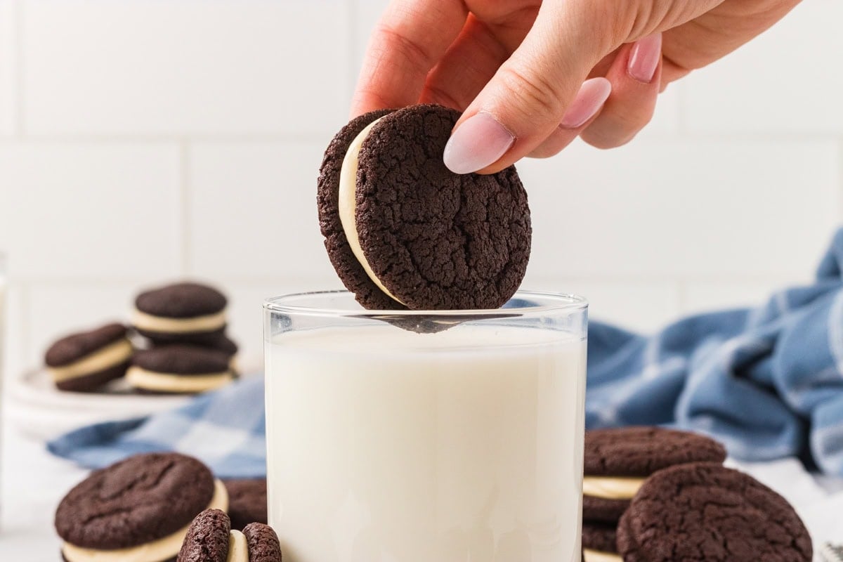 A hand dipping a homemade gluten free Oreo into a glass of milk, with more cookies and a blue checkered cloth in the background.