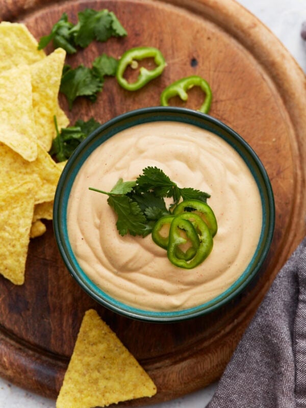 Bowl of creamy cashew queso dip topped with fresh cilantro and sliced jalapeños, surrounded by tortilla chips and cilantro on a wooden board.