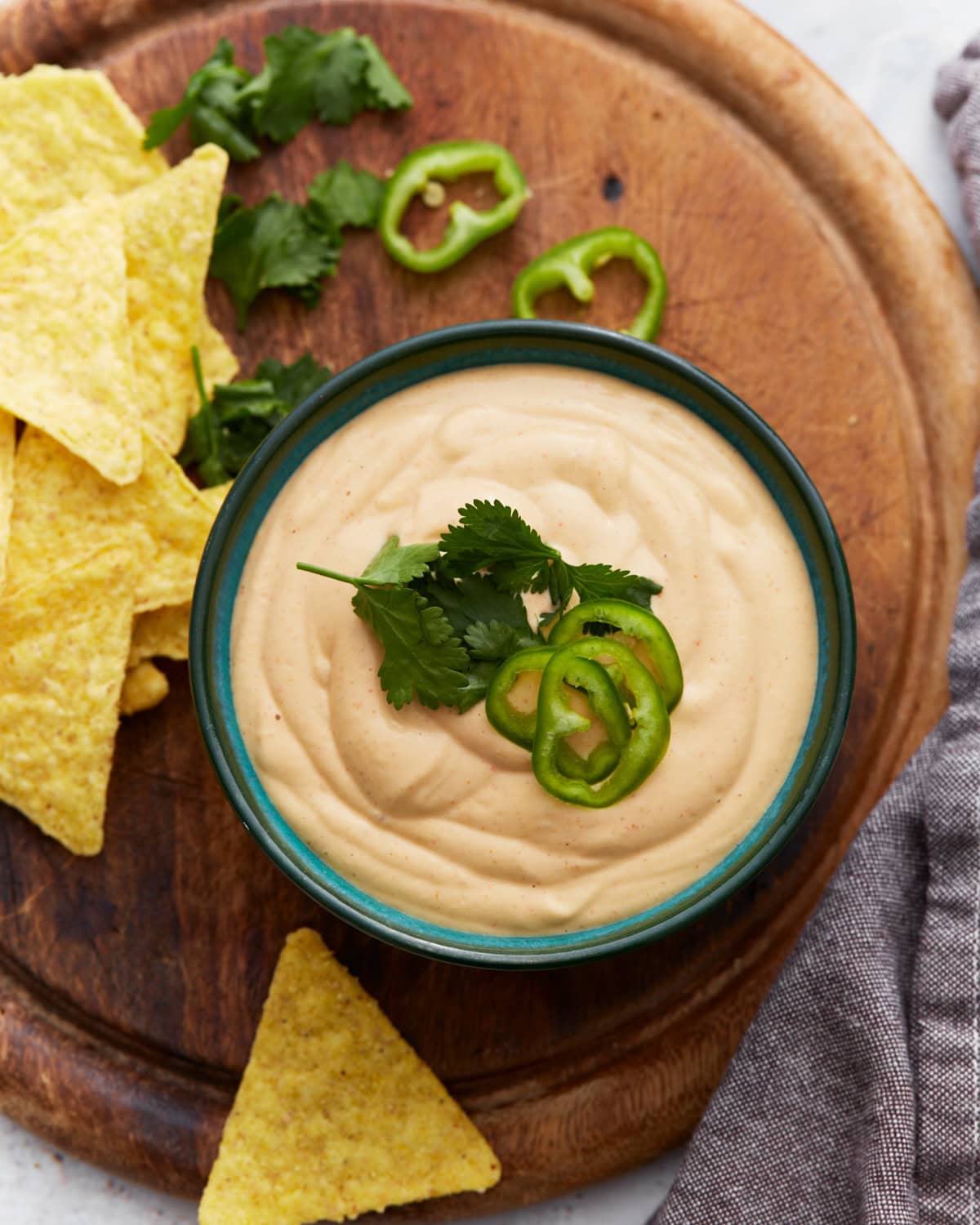 Bowl of creamy cashew queso dip topped with fresh cilantro and sliced jalapeños, surrounded by tortilla chips and cilantro on a wooden board.