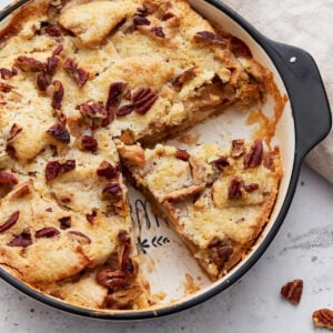 Overhead shot of a baked crustless apple pie in a round black-handled baking dish with one slice removed, showing the soft, golden topping and chopped pecans.