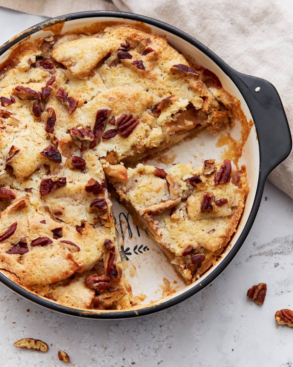 Overhead shot of a baked crustless apple pie in a round black-handled baking dish with one slice removed, showing the soft, golden topping and chopped pecans.