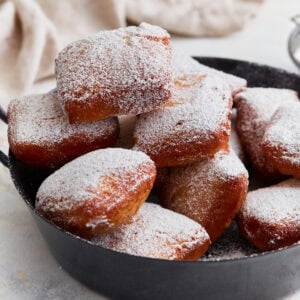 A pile of golden brown gluten-free beignets dusted with powdered sugar, served in a black pan.
