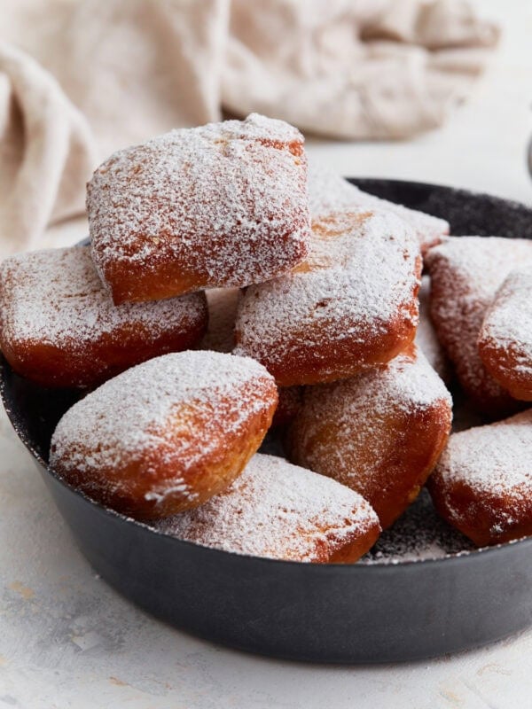 A pile of golden brown gluten-free beignets dusted with powdered sugar, served in a black pan.