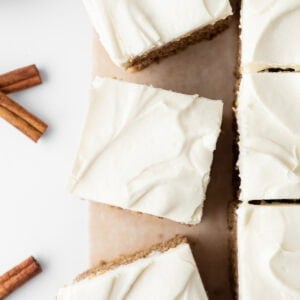 Top-down view of sliced gluten-free spice cake with cream cheese frosting on parchment paper, with cinnamon sticks nearby.
