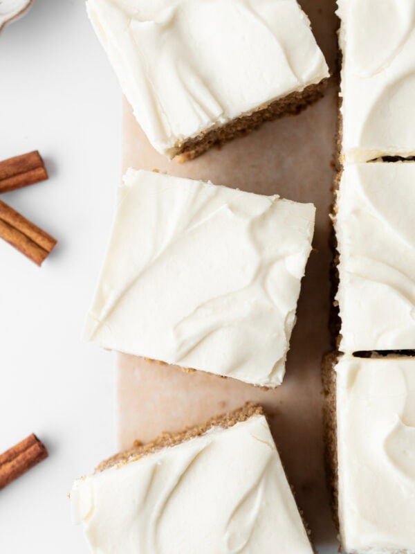 Top-down view of sliced gluten-free spice cake with cream cheese frosting on parchment paper, with cinnamon sticks nearby.