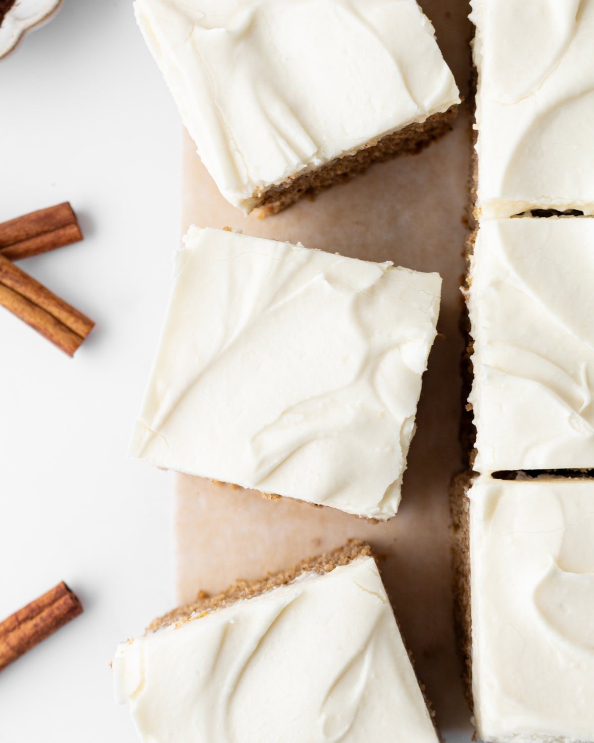 Top-down view of sliced gluten-free spice cake with cream cheese frosting on parchment paper, with cinnamon sticks nearby.