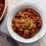 A white bowl filled with keto chili topped with shredded cheddar cheese, placed on a light surface with cheese scattered around.