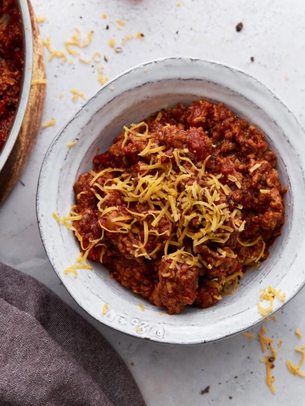 A white bowl filled with keto chili topped with shredded cheddar cheese, placed on a light surface with cheese scattered around.
