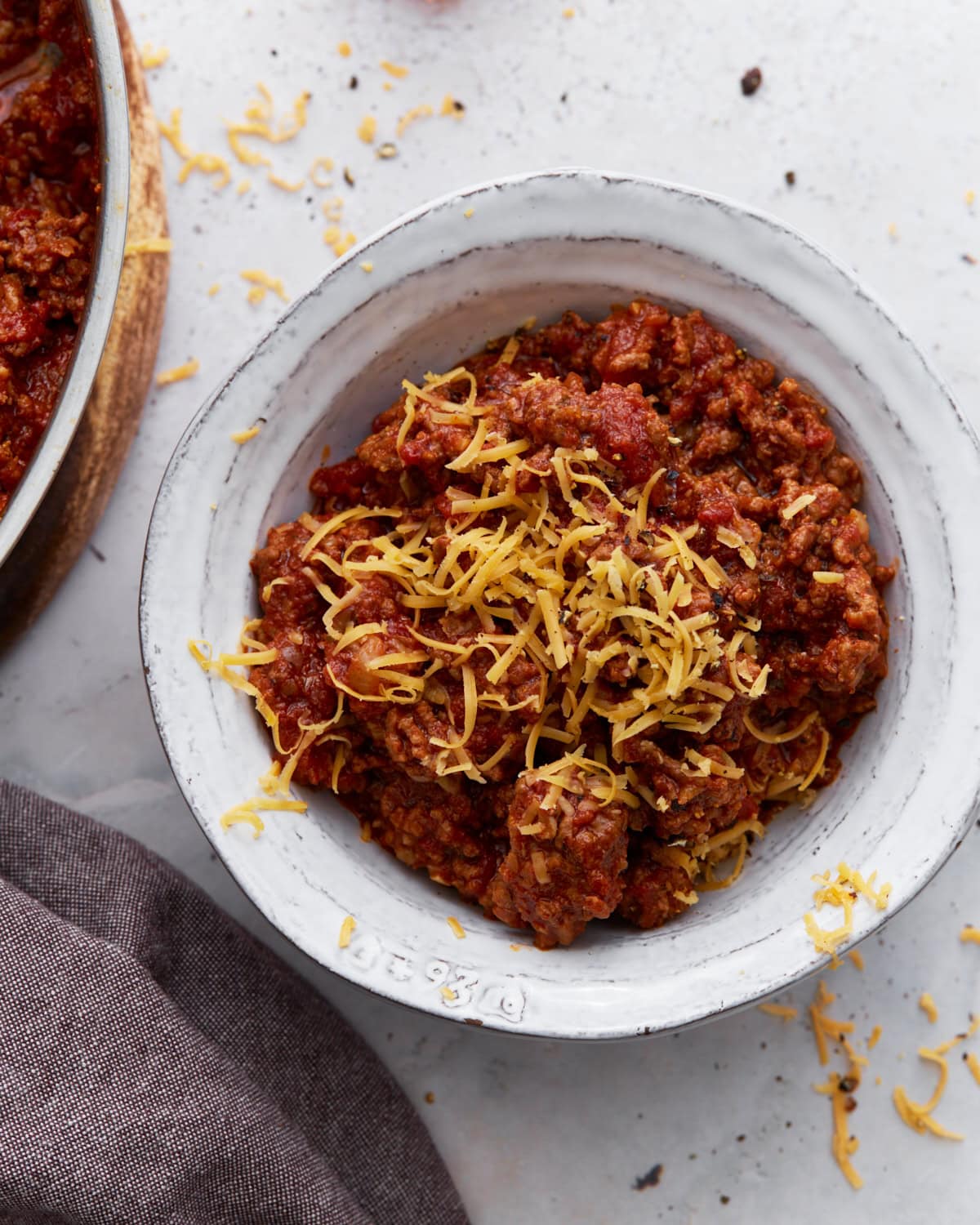 A white bowl filled with keto chili topped with shredded cheddar cheese, placed on a light surface with cheese scattered around.