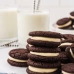 Gluten free chocolate sandwich cookies stacked on a wire rack, filled with vanilla marshmallow cream, with glasses of milk and striped straws in the background.