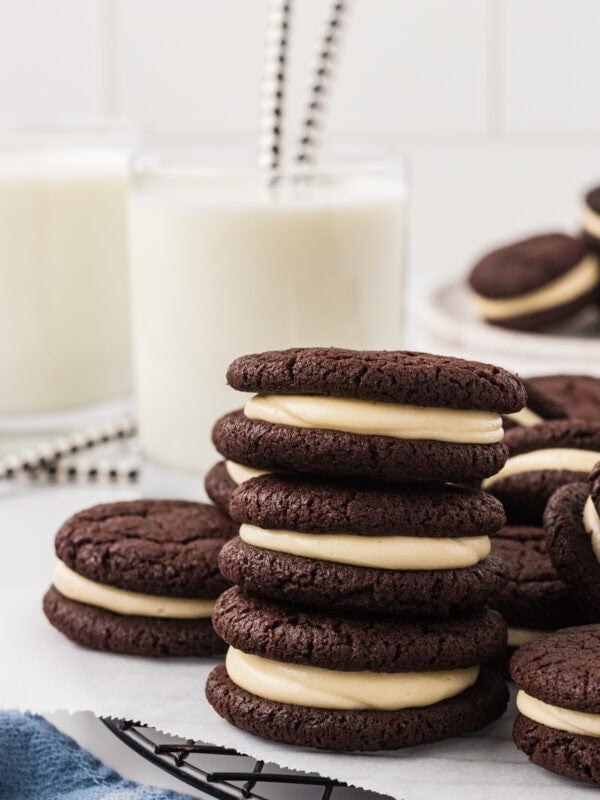 Gluten free chocolate sandwich cookies stacked on a wire rack, filled with vanilla marshmallow cream, with glasses of milk and striped straws in the background.