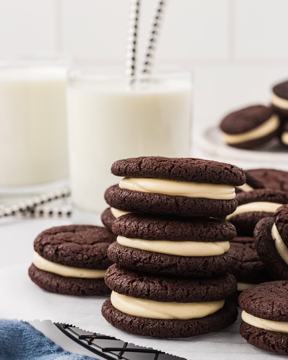 Gluten free chocolate sandwich cookies stacked on a wire rack, filled with vanilla marshmallow cream, with glasses of milk and striped straws in the background.