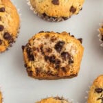 A close-up of gluten-free chocolate chip muffins on a white background, with one muffin cut in half to show the soft, fluffy interior and melty chocolate chips.