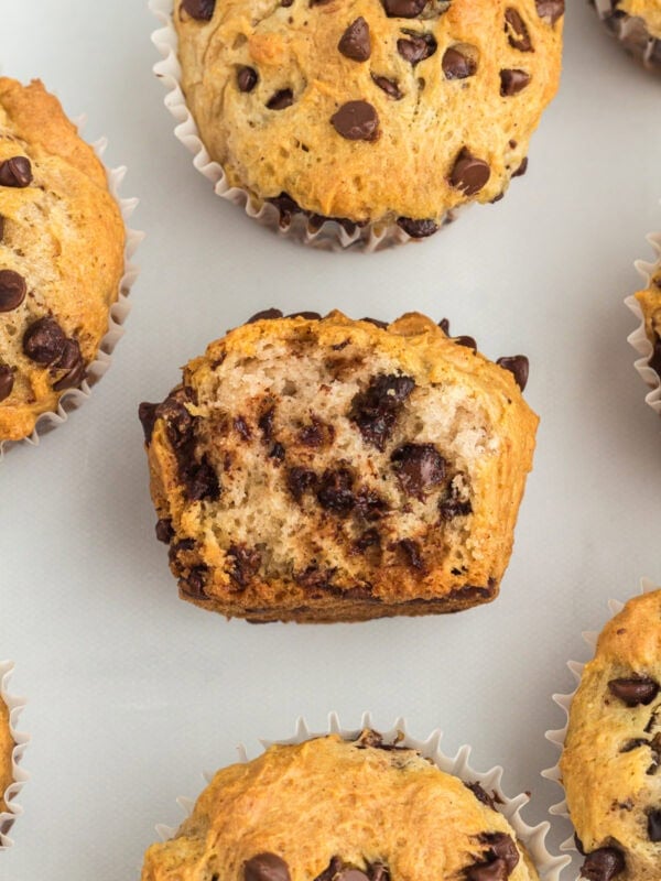 A close-up of gluten-free chocolate chip muffins on a white background, with one muffin cut in half to show the soft, fluffy interior and melty chocolate chips.