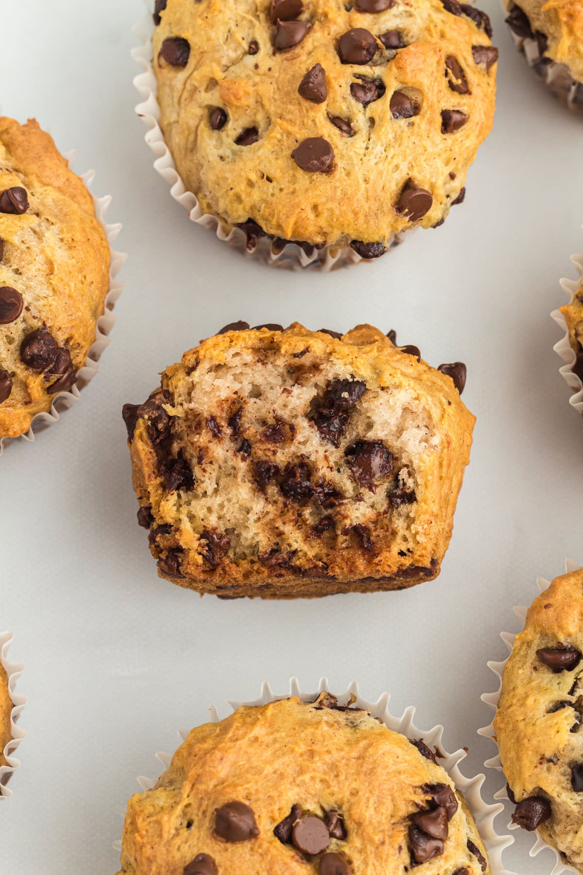 A close-up of gluten-free chocolate chip muffins on a white background, with one muffin cut in half to show the soft, fluffy interior and melty chocolate chips.