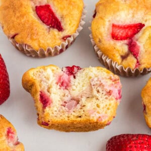 Gluten-free strawberry muffins with golden tops, one muffin cut in half to show the moist, fluffy crumb and chunks of fresh strawberries inside.