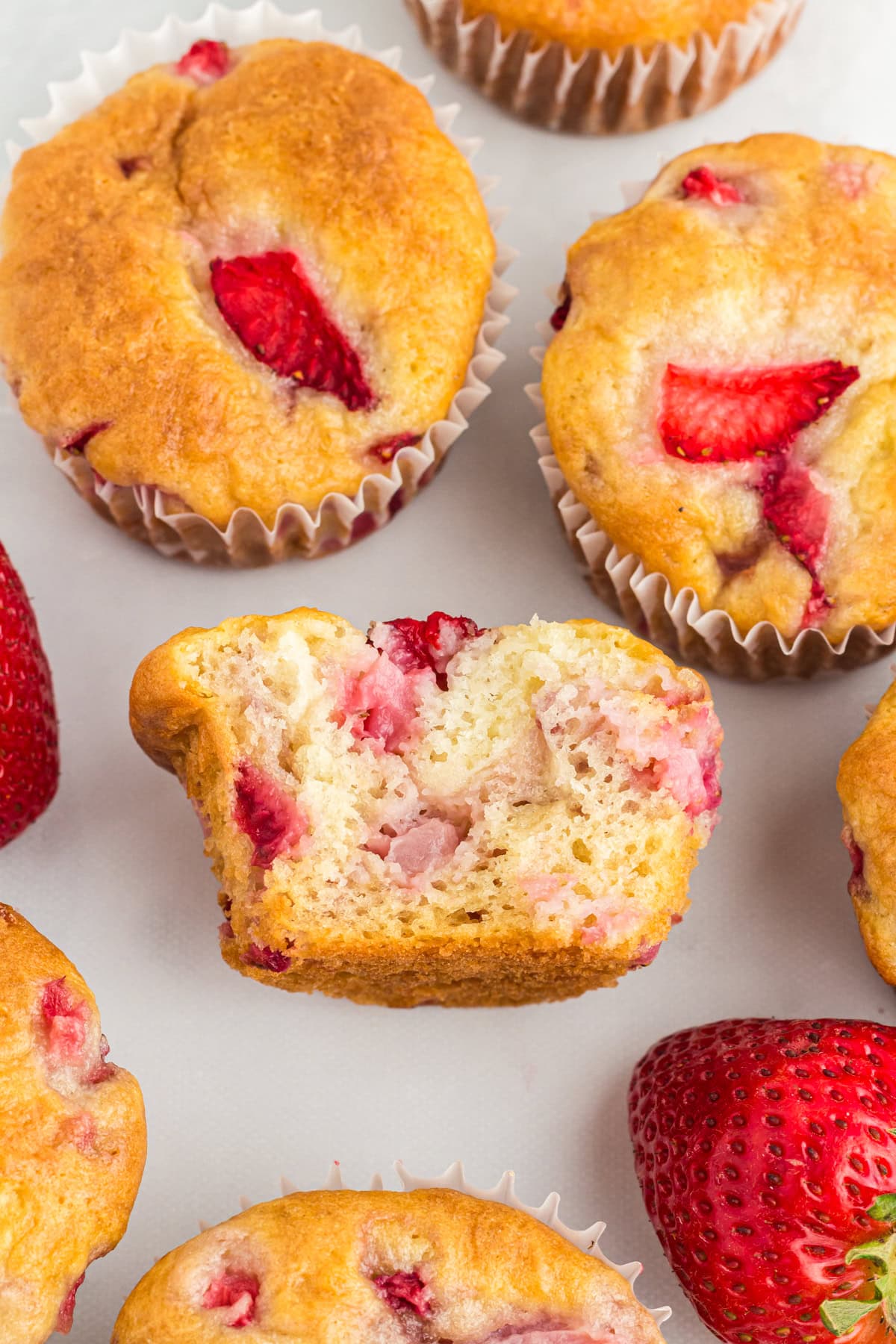 Gluten-free strawberry muffins with golden tops, one muffin cut in half to show the moist, fluffy crumb and chunks of fresh strawberries inside.