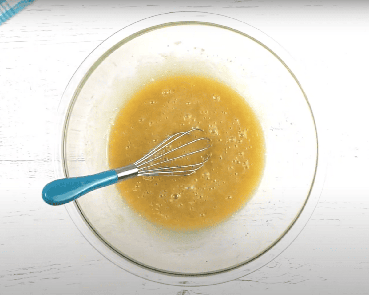 Whisked wet ingredients in a clear glass bowl with a blue-handled whisk, ready for mixing into muffin batter.