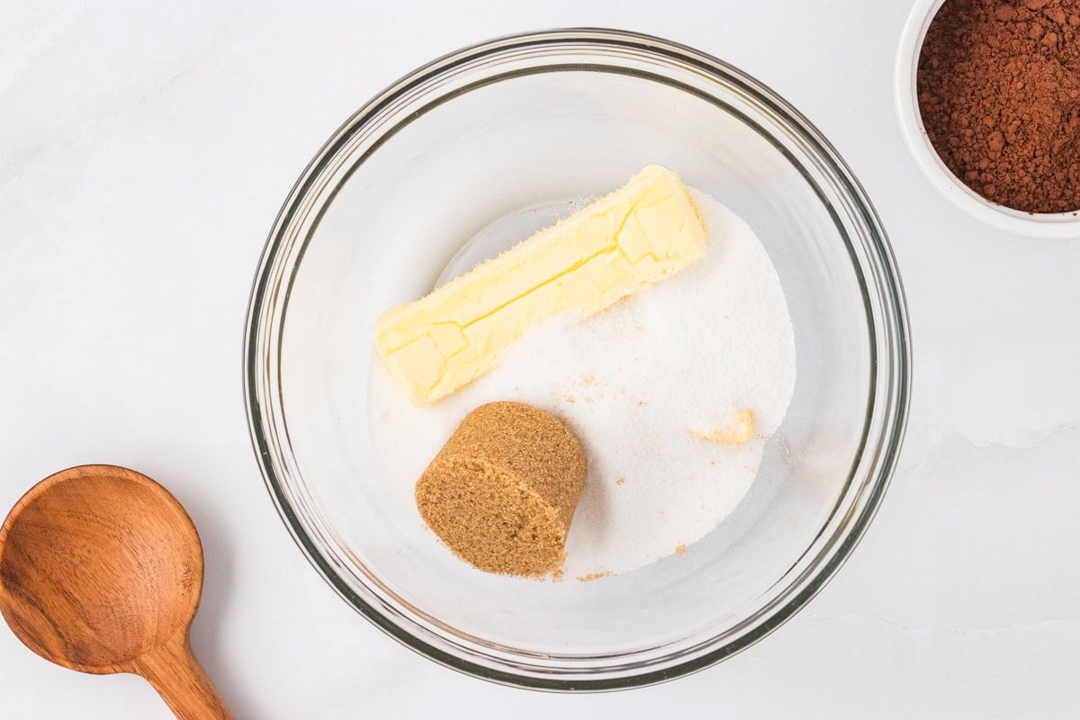 A glass mixing bowl with a stick of butter, granulated sugar, and packed brown sugar, ready to be creamed for cookie dough.