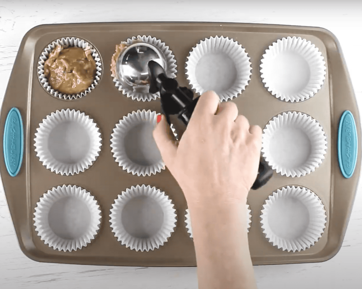 Muffin batter being scooped into a lined muffin tin using a spring-loaded scoop.