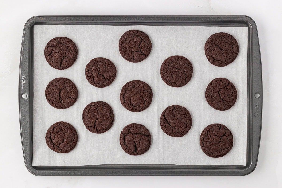 Freshly baked chocolate cookies cooling on a parchment-lined baking sheet with cracked tops and a rich color.