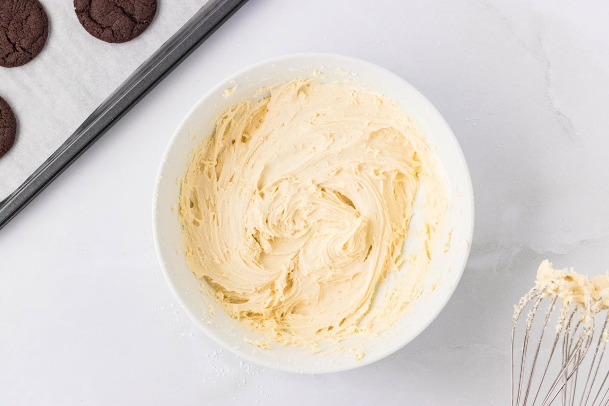 A white bowl filled with fluffy vanilla marshmallow filling, next to baked cookies and a whisk.