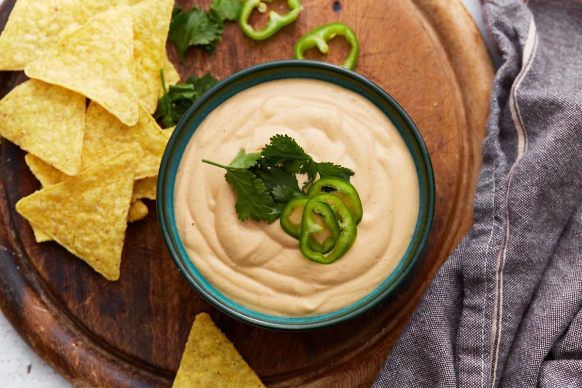 Bowl of cashew queso dip garnished with cilantro and jalapeño slices, next to tortilla chips on a wooden board with a gray napkin.