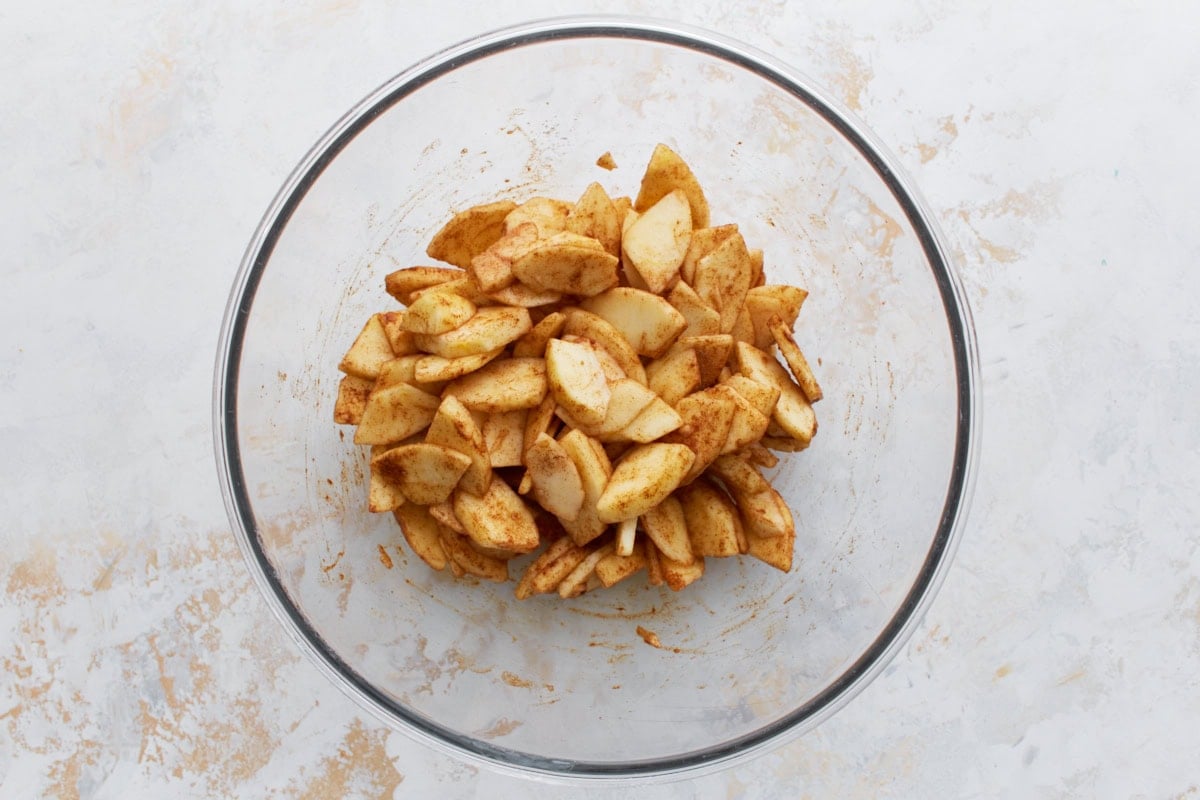 Sliced apples tossed with cinnamon, sugar, and lemon juice in a large clear glass mixing bowl.
