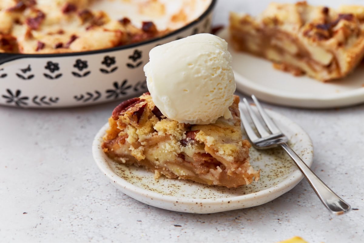 Slice of crustless apple pie topped with a scoop of vanilla ice cream on a small plate with a fork, with the full pie in the background.