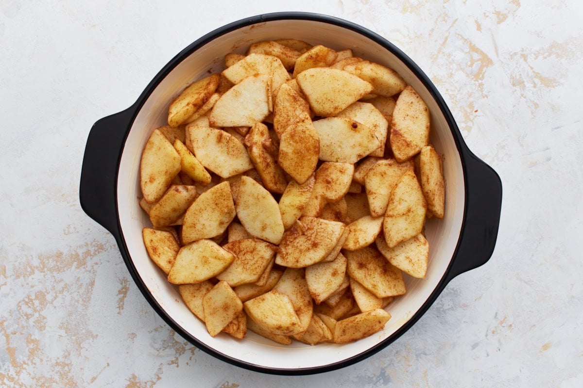 Seasoned apple slices arranged evenly in the bottom of a round black-handled baking dish.