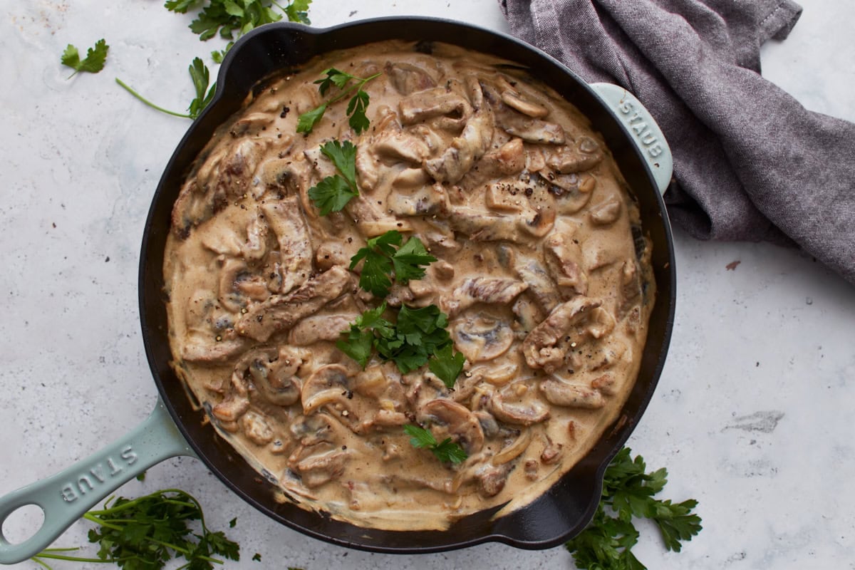 Gluten-free beef stroganoff served in a skillet and garnished with fresh parsley, placed on a white background with scattered herbs and a linen napkin.
