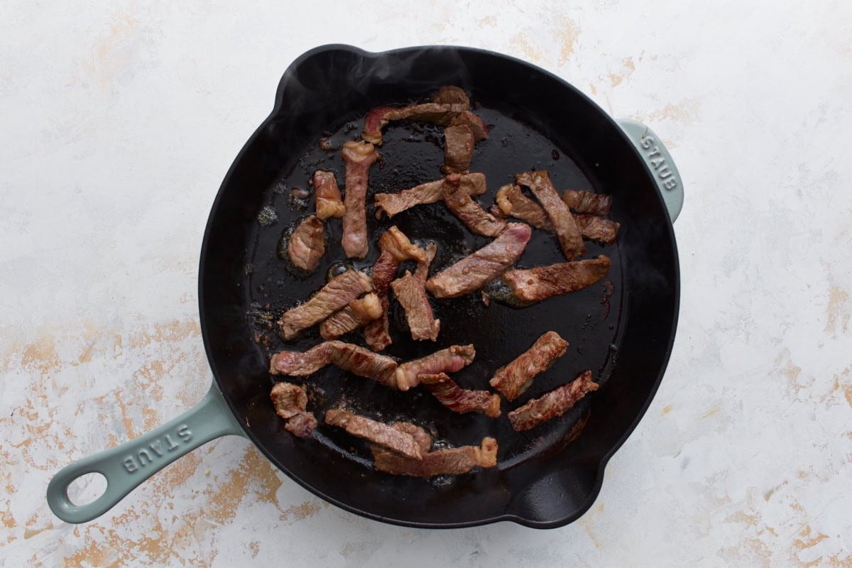 Beef strips searing in a hot cast iron skillet until browned.