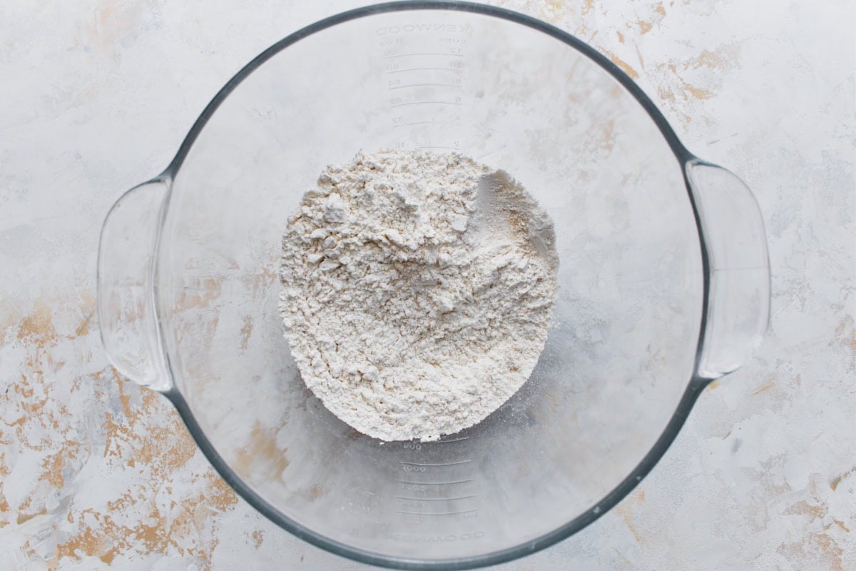 A glass mixing bowl filled with a blend of gluten-free flour, yeast, sugar, and salt.