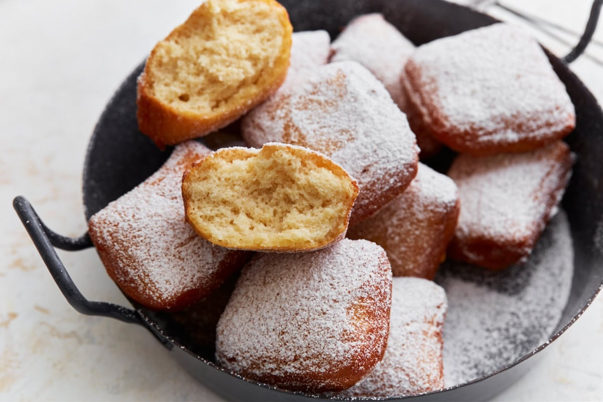 A close-up of a halved gluten-free beignet showing its fluffy interior, stacked on top of more powdered sugar–coated beignets in a black serving dish.