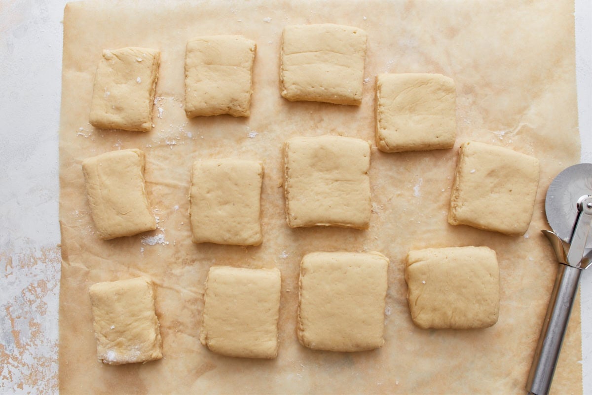 Squares of gluten-free beignet dough cut and spaced on parchment paper with a pizza cutter nearby.