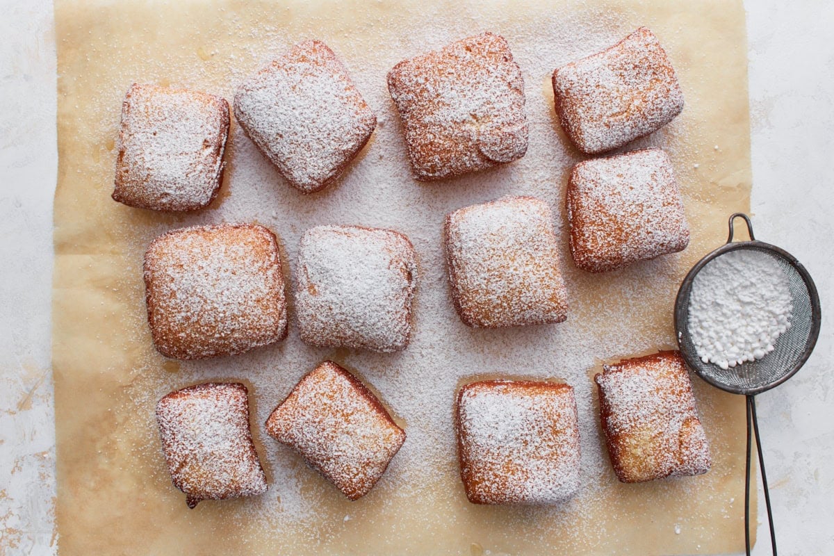 Freshly fried gluten-free beignets dusted with powdered sugar, arranged on parchment paper with a mesh sifter on the side.