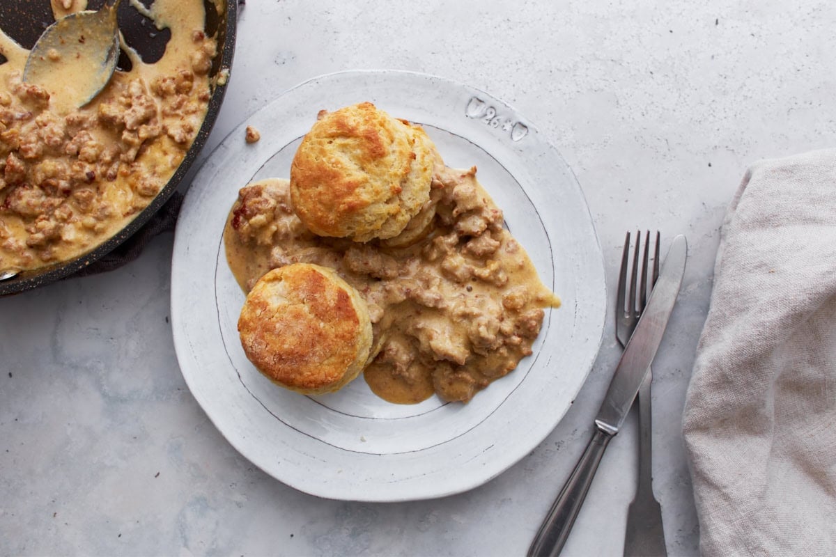 A plate with two golden biscuits covered in sausage gravy, served next to a pan of extra gravy, with a fork and knife on the side.